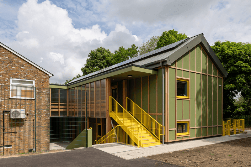 Modernes Gebäude mit grüner und gelber Fassade, mit großen Glasscheiben und einer Treppe vor einem bewölkten Himmel.