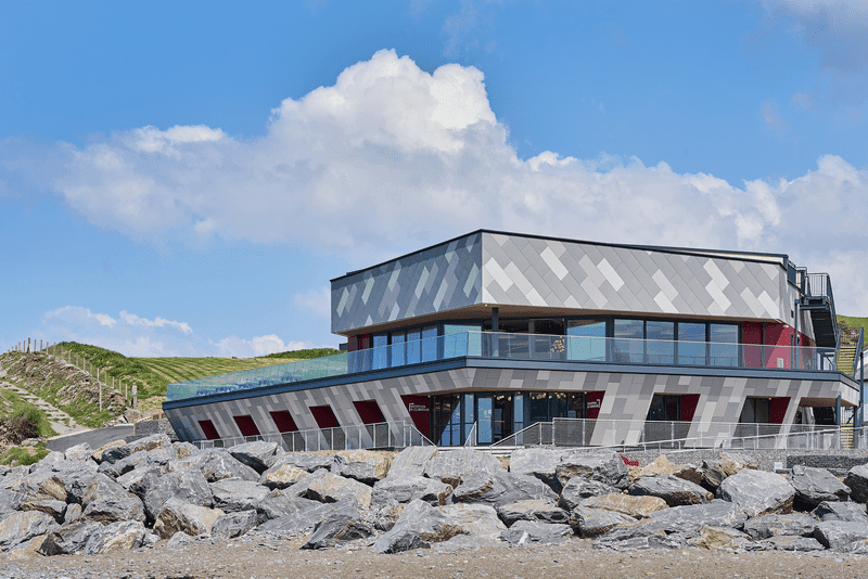 Modernes Gebäude am felsigen Ufer mit Flachdach und großen Glasfenstern vor einem blauen Himmel mit vereinzelten Wolken.