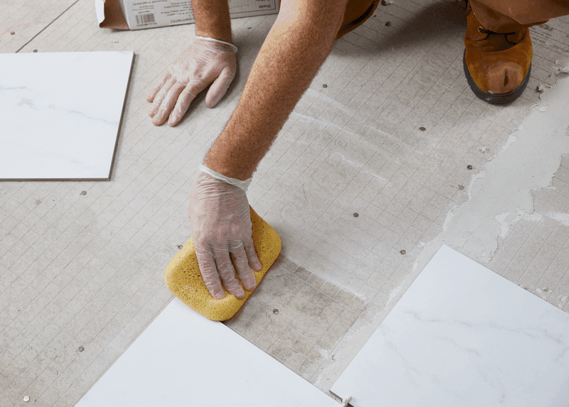 A person wearing gloves uses a sponge to clean grout between marble tiles on a floor.