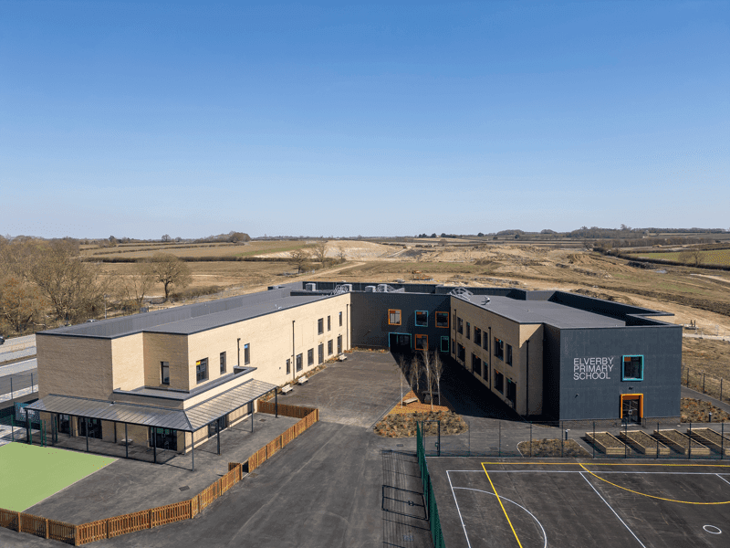 Luftaufnahme eines modernen Schulgebäudes, umgeben von einem offenen Feld und leeren Flächen. Das Gebäude besteht aus mehreren Abschnitten und einem Spielplatzbereich.