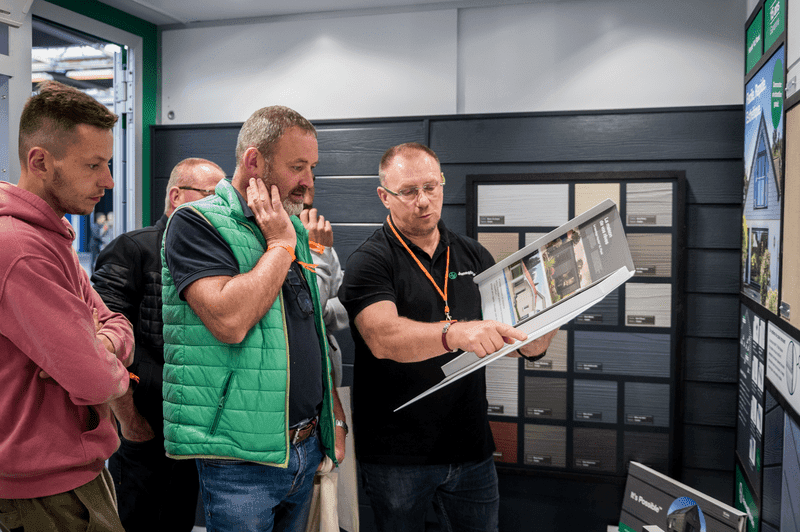 Eine Gruppe von Männern begutachtet eine Auslage, während ein Mann auf eine Broschüre zeigt und in einem gut beleuchteten Innenraum über die Möglichkeiten diskutiert.