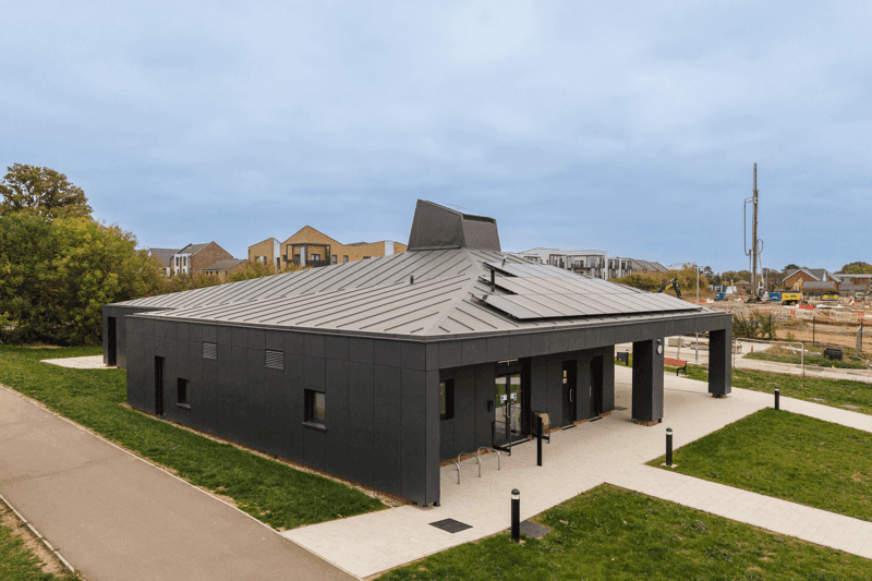 A modern, dark-colored building with a unique roof design, surrounded by grass and a pathway.