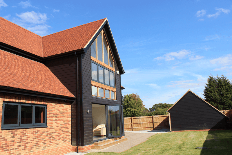A modern two-story house with a dark wooden exterior, large windows, and a red roof, set in a grassy yard under a blue sky.